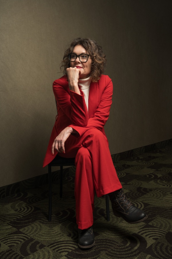 Tracy in a red outfit, sitting on a chair in a hallway, with one hand near her chin.